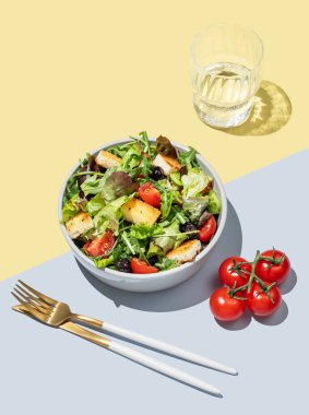 Marul, domates, kızarmış ekmek ve zeytinli salata sarı mavi bir arka planda, bir bardak su ve çatal bıçakla. Reklam menüsü veya afiş için sağlıklı diyet gıda konsepti. Üst görünüm, alanı kopyala.