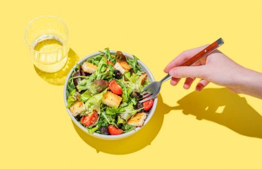 El ele tutuşan sebze salatası, marul, domates, kızarmış ekmek ve zeytin ile sarı arka planda bir bardak su ile birlikte. Reklam menüsü veya afiş için sağlıklı diyet gıda konsepti. Üst görünüm, alanı kopyala.