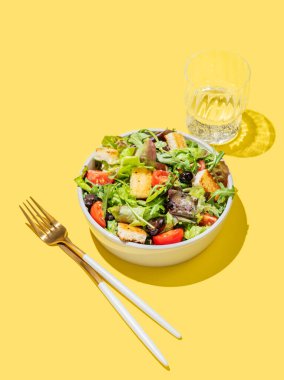 Sebzeli salata, marul, domates, kızarmış ekmek ve zeytinler sarı arka planda bir kasede, bir bardak su ve çatal bıçakla. Reklam menüsü veya afiş için sağlıklı diyet gıda konsepti. Boşluğu kopyala.