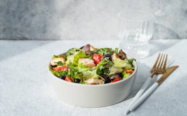 Salata, salata, domates, kızarmış ekmek, zeytin ve parmesan peyniri. Arka planda güneş ışığı, bir bardak su ve çatal bıçak. Sağlıklı beslenme konsepti. Ön görünüm, boşluğu kopyala.