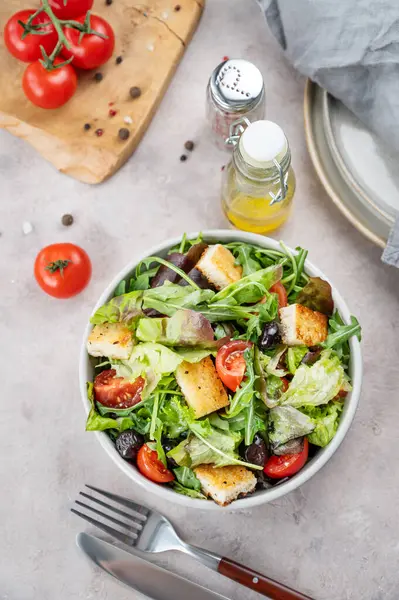 Baharat, yağ ve çatal bıçaklı hafif bir arka planda bir kasede marul, domates, kızarmış ekmek ve zeytinli sebze salatası. Sağlıklı beslenme konsepti. Üst görünüm.