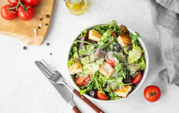 Marul, domates, kızarmış ekmek, zeytin ve parmesan peynirli vejetaryen salatası hafif bir arka planda taze sebze, yağ ve çatal bıçakla birlikte. Sağlıklı beslenme konsepti. Üst görünüm, alanı kopyala.