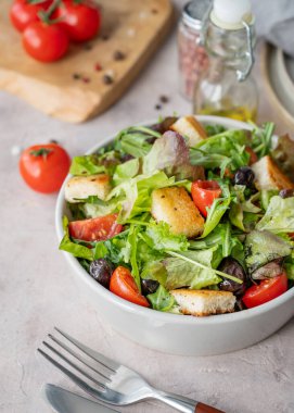 Baharat, yağ ve çatal bıçakla hafif arka planda bir kasede marul, domates, kızarmış ekmek ve zeytinli sebze salatası. Sağlıklı beslenme konsepti. 
