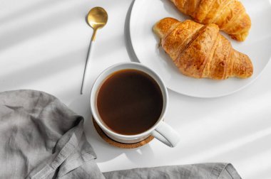 Kahve fincanı ve beyaz arka planda sabah gölgesi ve peçeteli taze kruvasanlar. Yaratıcı tasarım ve sağlıklı yemek ve Fransız kahvaltısı konsepti. Üst görünüm ve kopyalama alanı.