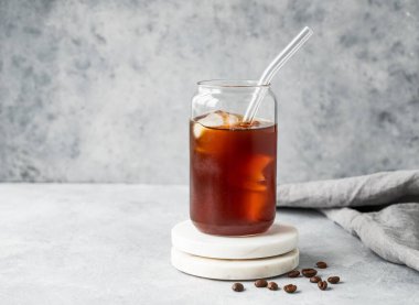 Kahve çekirdekleri ve peçeteyle birlikte açık beton arka plandaki mermer tezgahta buzlu soğuk demlenmiş kahve bardağı. Moda yazlık ferahlatıcı içecek. Ön görünüm, boşluğu kopyala.