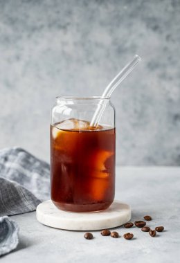 Kahve çekirdekleri ve peçeteyle birlikte açık beton arka plandaki mermer tezgahta buzlu soğuk demlenmiş kahve bardağı. Moda yazlık ferahlatıcı içecek. Ön görünüm, boşluğu kopyala.