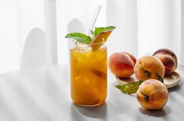 Şeftalili buzlu çay, cam kenarındaki beyaz masada taze meyve ve gölgeli bir bardakta nane. Bar ya da kafe menüsü için sağlıklı bir yaz içeceği konsepti. Ön görünüm, boşluğu kopyala.