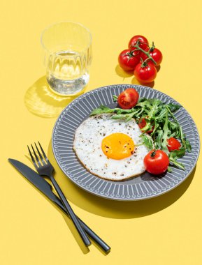 Koyu bir tabakta roka ve kiraz domatesli sahanda yumurta ve bir bardak su. Reklam menüsü ya da afiş kafe için sağlıklı kahvaltı konsepti. Boşluğu kopyala.