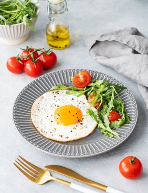 Koyu bir tabakta roka ve vişneli domatesli sahanda yumurta. Arka planda yemek, peçete ve yağ var. Kırsal tarzda sağlıklı, hızlı kahvaltı konsepti. Üst görünüm.
