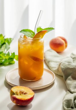Şeftalili buzlu çay, cam kenarındaki beyaz masada bardakta nane şekeri taze meyve, bitki ve gölgelerle birlikte. Bar ya da kafe menüsü için sağlıklı bir yaz içeceği konsepti. Ön görünüm, boşluğu kopyala.