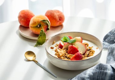 Doğal Yunan yoğurdu, granola veya müsli ve şeftali, kaşık, peçete ve sabah gölgeleriyle beyaz bir masada. Sağlıklı ve besleyici kahvaltı konsepti. Boşluğu kopyala.