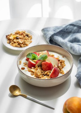 Doğal Yunan yoğurdu, granola veya müsli ve şeftali, kaşık, peçete ve sabah gölgeleriyle beyaz bir masada. Sağlıklı ve besleyici kahvaltı konsepti. Boşluğu kopyala.