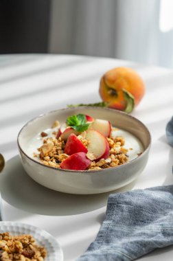 Doğal Yunan yoğurdu, granola veya müsli ve şeftali, peçeteli ve sabah gölgeli beyaz bir masada. Sağlıklı ve besleyici kahvaltı konsepti. Boşluğu kopyala.