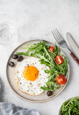 Arugula ile kızarmış yumurta, yumurtalar ve hafif arka planda bir tabakta vişne domatesleri, bir bardak su ve yemek ile birlikte. Kırsal tarzda sağlıklı, hızlı kahvaltı konsepti. Üst görünüm, alanı kopyala.