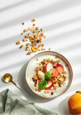 Beyaz arka planda şeftali, nane ve dağınık granola kaplı doğal Yunan yoğurdu, kaşık, taze meyve, peçete ve sabah gölgesi. Sağlıklı ve besleyici kahvaltı konsepti. Üst görünüm, alanı kopyala