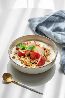 Doğal Yunan yoğurdu, granola veya müsli ve şeftali, kaşık, peçete ve sabah gölgeleriyle beyaz bir masada. Sağlıklı ve besleyici kahvaltı konsepti. Boşluğu kopyala.