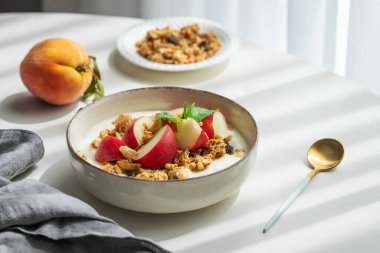 Doğal Yunan yoğurdu, granola veya müsli ve şeftali, kaşık, peçete ve sabah gölgeleriyle beyaz bir masada. Sağlıklı ve besleyici kahvaltı konsepti. Boşluğu kopyala.