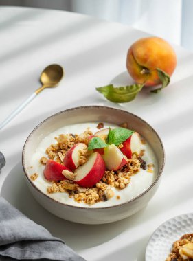 Doğal Yunan yoğurdu, granola ve şeftali, cam kenarındaki beyaz bir masada kaşık, peçete ve sabah gölgeleriyle. Sağlıklı ve besleyici kahvaltı konsepti. Üst görünüm, alanı kopyala.
