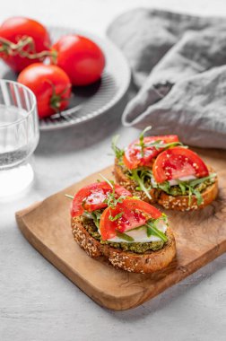 Domatesli sağlıklı susamlı tost, mozzarella, roka ve ahşap tahtada pesto hafif arka planda bir bardak su, taze sebze ve sabah ışığı. Kahvaltı atıştırmalığı konsepti.