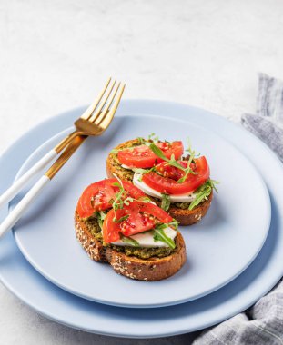 Domatesli sağlıklı susamlı tost, mozzarella, roka ve pesto hafif arka planda mavi bir tabakta, çatal bıçak ve peçeteyle. Kahvaltılık sandviç konsepti. Boşluğu kopyala.