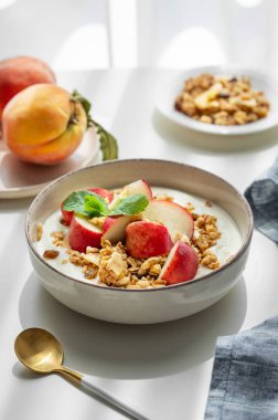 Doğal Yunan yoğurdu, granola ya da müsli ve şeftali bir kâse içinde beyaz bir masa üzerinde kaşık ve sabah gölgesi olan bir peçete. Sağlıklı ve besleyici kahvaltı konsepti. Boşluğu kopyala.