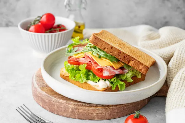 Domuz pastırmalı, peynirli, domatesli, marullu, roka ve soslu tost sandviçi, tahta bir tabakta, arka planda hafif bir peçeteyle. Sağlıklı ve besleyici kahvaltı konsepti. 