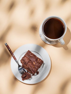 Bej arka planda bir tabakta çikolatalı kek, bir fincan taze demlenmiş kahve ve sabah gölgesi. Kafe menüsü reklamları ya da afişler için lezzetli hamur işleri kavramı. Üst görünüm, alanı kopyala.