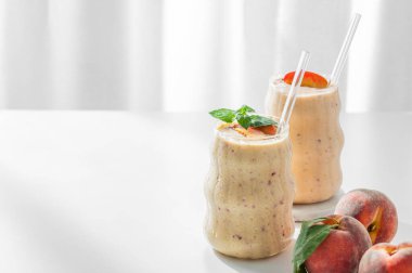 İki bardak şeftalili ve muzlu smoothie pencerenin yanında açık bir arka planda, tropikal meyveler ve sabah güneşiyle birlikte. Sağlıklı beslenme ve reklam menüsü kafesi ya da afişi için detoks diyeti kavramı. Ön görünüm, boşluğu kopyala.