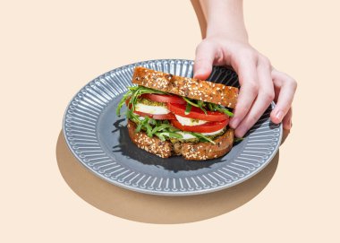 Elinde domatesli tahıllı ekmek, mozzarella roka ve pesto ile parlak bej arka planda gölgeli bir tabakta. Reklam menüsü ya da afiş için kahvaltılık tost konsepti. Boşluğu kopyala