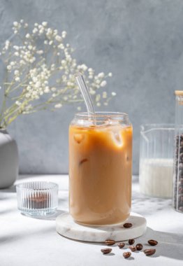 Bir bardak buzlu latte kahve, mermer tezgahın üzerinde süt ve çiçek buketi, kahve çekirdekleri ve sabah gölgeleri. Moda yazlık ferahlatıcı içecek konsepti. Ön görünüm, boşluğu kopyala.