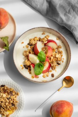 Doğal Yunan yoğurdu, granola veya müsli, şeftali ve nane, beyaz arka planda taze meyve, kaşık ve sabah gölgeleriyle dolu bir kasede. Sağlıklı ve besleyici kahvaltı konsepti. Üst görünüm