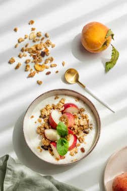 Beyaz arka planda şeftali, nane ve dağınık granola kaplı doğal Yunan yoğurdu, kaşık, taze meyve, peçete ve sabah gölgesi. Sağlıklı ve besleyici kahvaltı konsepti. Üst görünüm, alanı kopyala