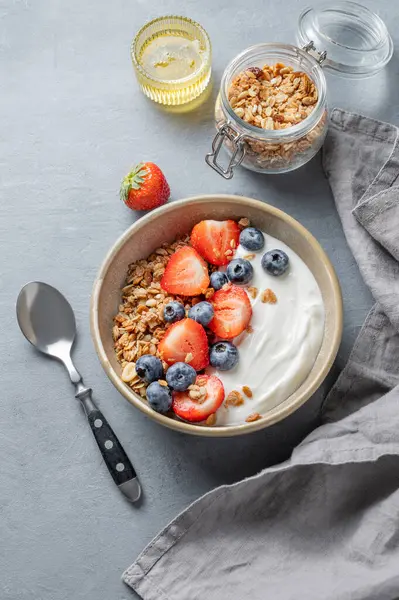 Mısır gevreği, yabanmersini ve çilekli yoğurt mavi arka planda bal, kaşık ve peçeteli bir kasede. Sağlıklı ve besleyici kahvaltı konsepti. Üst görünüm, alanı kopyala.