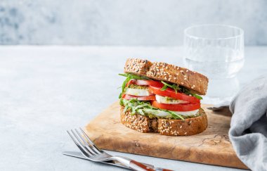 Taze domatesli tahıllı ekmek, mozzarella roka ve ahşap bir tahtada pesto. Arka planda hafif dokulu bir cam su ve çatal bıçakla. Sağlıklı beslenme ve beslenme kahvaltısı kavramı. Boşluğu kopyala.