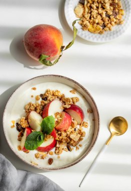 Doğal Yunan yoğurdu, granola veya müsli, şeftali ve nane karışımı beyaz bir arka planda, kaşık, peçete ve sabah gölgeleriyle. Sağlıklı ve besleyici kahvaltı konsepti. Üst görünüm, alanı kopyala