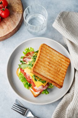 Domuz pastırmalı, peynirli, domatesli, marullu, roka soslu tost sandviçi mavi bir arka planda bir bardak su ve peçeteyle. Sağlıklı ve besleyici kahvaltı konsepti. Üst görünüm.