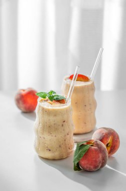 İki bardak şeftalili ve muzlu smoothie pencerenin yanında açık bir arka planda, tropikal meyveler ve sabah güneşiyle birlikte. Sağlıklı beslenme ve reklam menüsü kafesi ya da afişi için detoks diyeti kavramı. Ön görünüm, boşluğu kopyala.