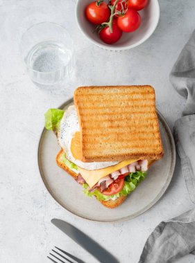 Yumurtalı, pastırmalı, peynirli, domatesli, marullu ve soslu tost sandviçi hafif bir arka planda, bir bardak su ve çatal bıçakla. Sağlıklı ve besleyici kahvaltı konsepti. Üst görünüm.