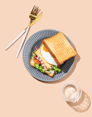 Yumurtalı, pastırmalı, peynirli, domatesli, salatalı ve soslu sandviç bej bir arka planda, bir bardak su ve çatal bıçaklı. Reklam menüsü ya da afiş kafe için sağlıklı ve besleyici tost kahvaltısı konsepti. Üst görünüm.