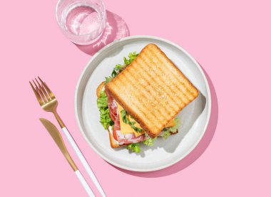 Pastırmalı, peynirli, domatesli, marullu, roka soslu ve pembe arka planda bir tabakta bir tost ekmeği, bir bardak suyla. Menü kafenin reklam afişi için sağlıklı ve besleyici kahvaltı konsepti. Üst görünüm.