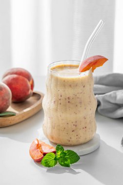 Şeftalili ve muzlu meyve püresi, cam kenarındaki beyaz bir masada, tropik meyve dilimleri ve sabah güneşiyle birlikte. Sağlıklı beslenme ve detoks diyeti kavramı. Ön görünüm, boşluğu kopyala.