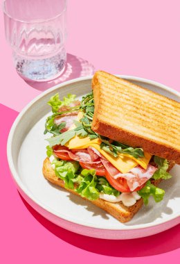 Domuz pastırmalı, peynirli, domatesli, marullu, roka soslu tost ekmeği. Pembe bir arka planda, bir bardak suyla birlikte. Menü kafenin reklam afişi için sağlıklı ve besleyici kahvaltı konsepti. Boşluğu kopyala.