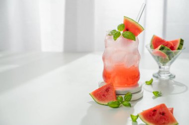 Buzlu karpuz mojito kokteyli ve bir bardak nane şekeri pencerenin yanındaki beyaz masada taze dilimler ve güneş ışığıyla. Bar menüsü ya da afiş için yaz ferahlatıcı limonata içeceği konsepti. Ön görünüm, boşluğu kopyala.