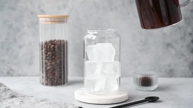 Soğuk kahve, mermer tezgahta buzlu bir bardağa kahve çekirdekleri ve kaşıkla birlikte hafif beton bir zemin üzerine dökülüyor. El yapımı moda yaz ferahlatıcı içecek. Ön görünüm.