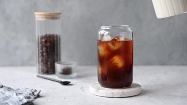 Süt sürahisinden bir bardak buzlu kahveye, açık fonda kahve çekirdekli mermer bir tahtayla dökülüyor. Moda yazlık ferahlatıcı içecek konsepti. Ön görünüm.