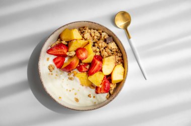 Mısır gevreği ya da müsli, doğal Yunan yoğurdu, çilek ve şeftali karışımı beyaz bir arka planda, kaşık ve sabah güneşiyle birlikte. Sağlıklı ve besleyici kahvaltı konsepti. Üst görünüm, alanı kopyala.