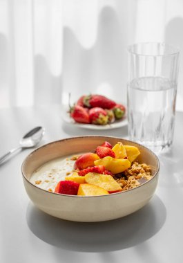 Doğal Yunan yoğurdu, çilek ve şeftalili granola ve müsli taze böğürtlen, bir bardak su ve sabah gün ışığıyla birlikte beyaz bir masada beyaz bir masada. Sağlıklı ve besleyici kahvaltı konsepti. Boşluğu kopyala.