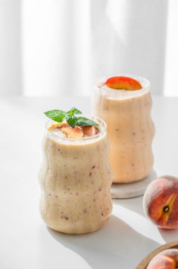 İki bardak şeftalili ve muzlu smoothie pencerenin yanında açık bir arka planda, tropikal meyveler ve sabah güneşiyle birlikte. Sağlıklı beslenme ve detoks diyeti kavramı. Ön görünüm, boşluğu kopyala.