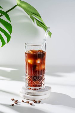 Hafif arka planda buzlu soğuk kahve bardağı, kahve çekirdekleri, canavar çiçeği yaprakları ve sabah gölgesi. Reklam menüsü kafe için yaz ferahlatıcı moda içecek konsepti. Ön görünüm, boşluğu kopyala.