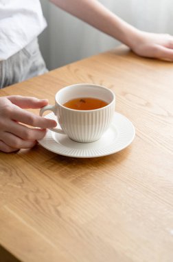 Sabah güneşli pencerenin yanındaki tahta masanın üstünde limonlu ve otlu bir fincan beyaz çay tutan kadın. Kahvaltı ve sağlıklı beslenme konsepti. Boşluğu kopyala.
